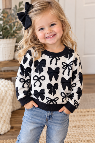 A young girl with blonde hair wearing a cream knit sweater with black bow patterns and blue jeans, standing in a cozy indoor setting.