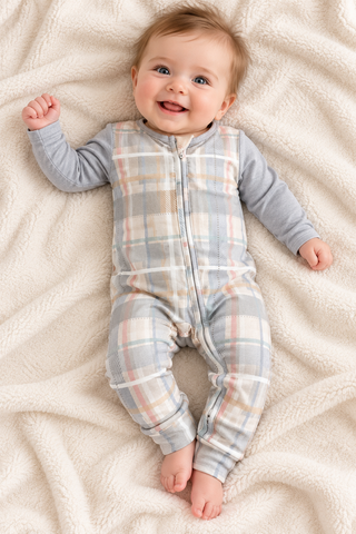 A baby lying on a soft cream blanket, wearing a long-sleeve zip-front footed sleeper with a light gray base and a soft plaid pattern in beige, white, and pale pink.
