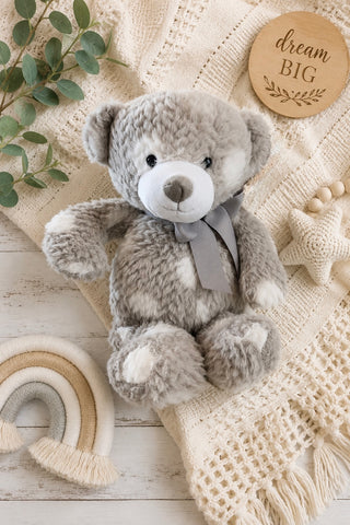 A plush gray and white teddy bear with a gray ribbon bow sits on a cream knit blanket with a wooden 'dream BIG' sign, eucalyptus leaves, a small rainbow decoration, and a star toy nearby.