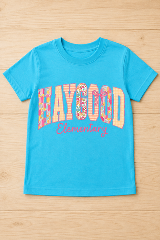 A bright turquoise blue short-sleeve t-shirt with 'HAYGOOD Elementary' printed across the chest in a floral patterned font, laid flat on a light wood background.