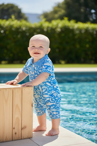 Baby in a blue swim outfit standing by a pool with greenery in the background