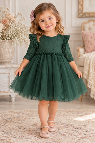 A young girl with curly light brown hair wearing a dark green tulle party dress with ruffled sleeves and a pink bow in her hair, standing on a patterned rug in a beautifully decorated room.