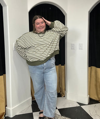 A woman modeling a green and white striped long-sleeve sweatshirt with an olive green hem, paired with light-wash blue jeans, standing in a fitting room with black and gold curtains.