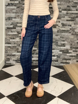 A woman modeling a cream ribbed long-sleeve mock neck sweater and dark blue plaid wide-leg jeans, standing in front of a gray stone tile wall and a black and white checkered floor.