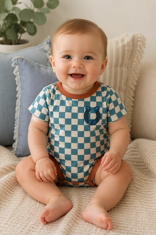 A baby sitting on a textured beige blanket in front of pillows, wearing a short-sleeve bodysuit with a blue and white checkered pattern and a blue horseshoe graphic on the chest.
