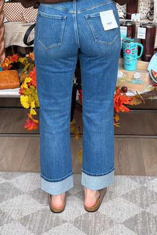 A person shown from the back wearing medium-wash blue cuffed jeans with a white brand tag visible in the back pocket, standing indoors on a patterned rug near a table with fall decorations and mugs.