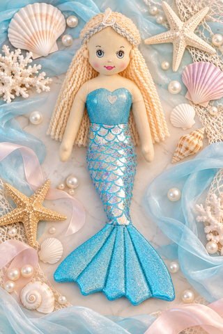 A mermaid doll with long blonde braided hair, a blue glittery scale tail, and a matching blue tail fin, styled on a light marble surface with seashells, starfish, pearls, coral, and sheer blue ribbon.