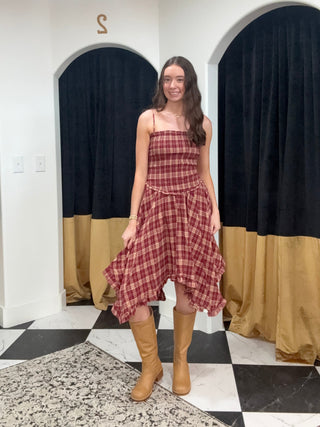A woman stands in a fitting room wearing a red and brown plaid strapless dress with a ruffled hem and tan knee-high boots. The background features white walls, arched fitting cubicles with black and gold curtains, and a black-and-white checkered floor.