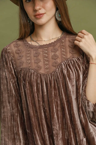 A close-up of a woman wearing a brown velvet long-sleeve top with a sheer embroidered yoke and gathered neckline, against an olive green background.