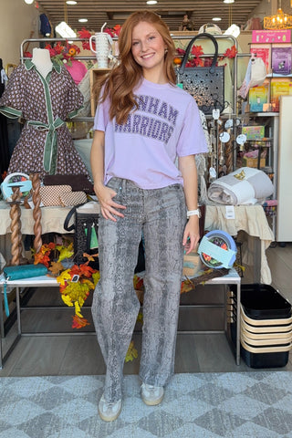 A woman stands in a boutique wearing a light lavender graphic t-shirt with 'BONHAM WARRIORS' in polkadots, paired with gray snakeskin-patterned jeans and white sneakers.