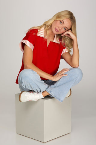 Woman in a red top and blue jeans sitting on a white cube against a plain background