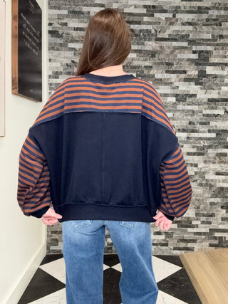 A person viewed from behind wearing a colorblock sweatshirt with a navy blue body, rust-orange and navy horizontal stripes on the shoulders and sleeves, and blue jeans, standing in front of a gray stone tile wall.