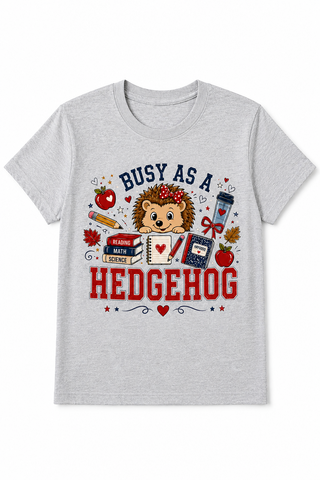 A light heather gray short-sleeve t-shirt with a colorful graphic of a cartoon hedgehog surrounded by school and nature-themed items, with the text 'BUSY AS A HEDGEHOG' printed on the front.
