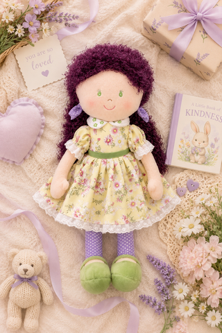 A soft plush doll with curly dark purple hair, a yellow floral dress, and green shoes is arranged on a cream fabric surface with flowers, a teddy bear, a gift box, and a small book around it.
