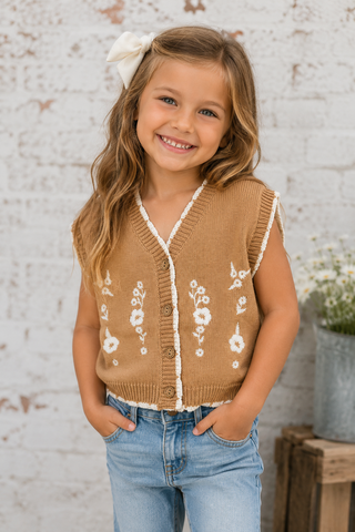 A young girl with a white bow in her hair is smiling, wearing a sleeveless tan knit vest with white floral embroidery and button details, paired with blue jeans, set against a rustic white brick wall background.