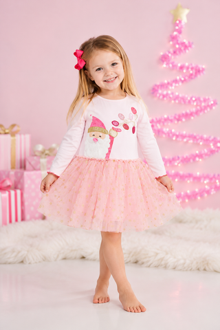 A young girl stands barefoot in a pink studio setting wearing a white long-sleeve top with a festive Santa graphic and a light pink tulle tutu skirt. She has a pink bow in her hair, with pink gift boxes and a glowing pink Christmas tree in the background.