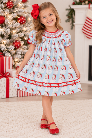 A young girl wearing a light blue Christmas-themed dress with a santa pattern and red trim, standing in a decorated holiday room with a Christmas tree, wrapped gifts, and a fireplace.