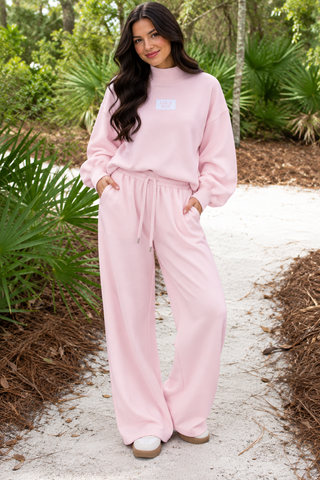 A woman stands on a sandy path outdoors wearing a light pink lounge set consisting of a sweatshirt and matching wide-leg pants. The set has a small white rectangular logo patch on the chest.