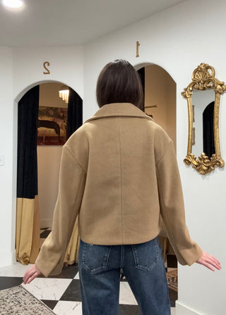 A person viewed from behind wearing a camel-colored long-sleeve jacket and blue jeans, standing in an indoor room with white walls, arches, a gold mirror, and a checkered floor.