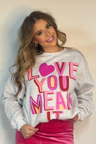 Woman wearing a gray sweatshirt with colorful text, pink skirt, and white boots against a plain background