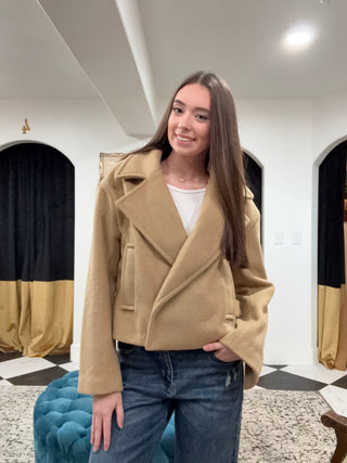 A young woman wearing a camel-colored suede moto jacket over a white t-shirt and blue jeans, standing in an indoor room with a black and gold tufted bench in the foreground.