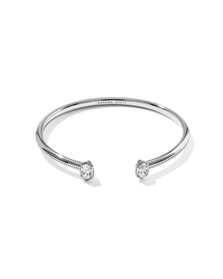 A silver open cuff bangle bracelet with two bezel-set oval clear gemstones, shown on a clean white background. The brand name 'KENDRA SCOTT' is engraved on the inside of the band.