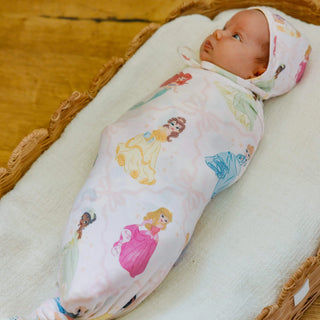 Newborn baby wrapped in a princess-themed swaddle blanket on a wooden surface.
