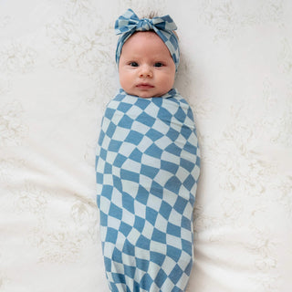 Baby swaddled in a blue checkered blanket with a matching headband on a white background