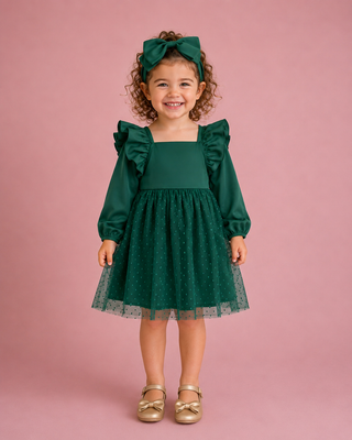 A young girl with curly brown hair wearing a dark green long-sleeve dress with ruffled shoulders, a matching green bow headband, and gold shoes, standing against a solid pink studio background.