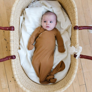 Baby in a brown knotted gown lying in a moses basket on a wooden floor.