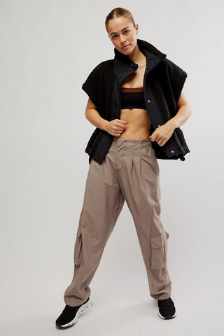 A model posing in a studio setting wearing a black fleece vest, a dark brown sports bra, loose taupe cargo pants, and black sneakers.