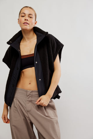 A model wearing a black fleece-lined zip-up vest over a brown sports bra and taupe trousers, photographed against a neutral light gray studio background.