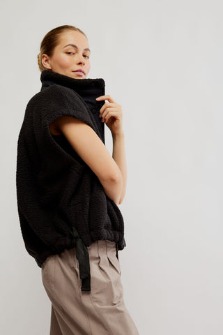 A woman wearing a black textured sleeveless mock neck vest with a drawstring tie, paired with beige trousers, photographed against a solid light gray background.