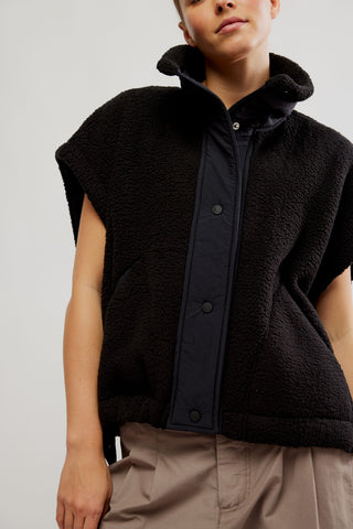 A close-up of a model wearing a black sherpa fleece sleeveless vest with a dark navy button-front placket and a high collar, paired with beige trousers.