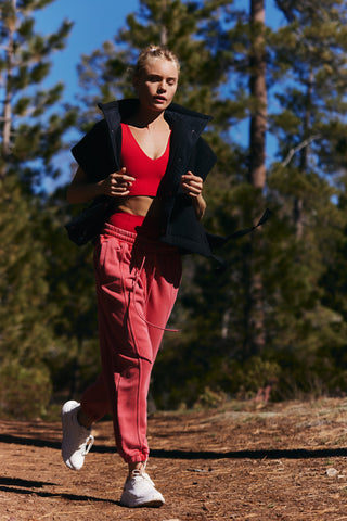 A woman jogging outdoors on a dirt path surrounded by pine trees, wearing a red sports bra, pink jogger pants, a black zip-up jacket, and white sneakers.