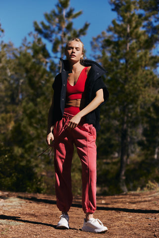 A woman stands outdoors on a dirt path among pine trees, wearing a red sports bra, coral pink jogger pants, a black open-front jacket, and white sneakers.