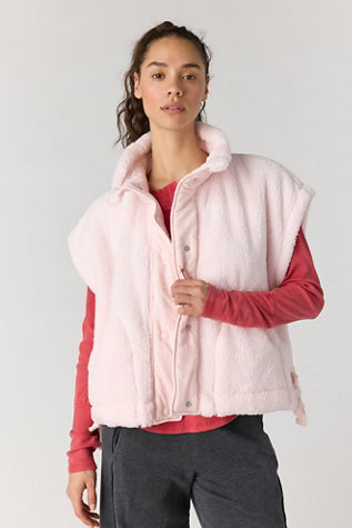 A woman modeling a light pink fuzzy sleeveless puffer vest over a coral pink long-sleeve shirt and dark gray pants, photographed against a light gray studio background.