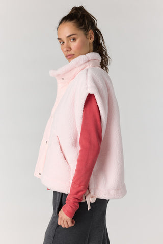 A woman wearing a light pink fuzzy sleeveless vest over a coral pink long-sleeve top and dark gray pants, photographed against a plain light gray studio background.