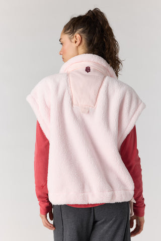A woman shown from the back wearing a pale pink sherpa fleece vest over a red long-sleeve top and dark gray pants, against a light gray studio background.