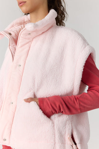 A close-up of a person wearing a light pink sherpa fleece vest over a red long-sleeve top, against a plain light gray background.