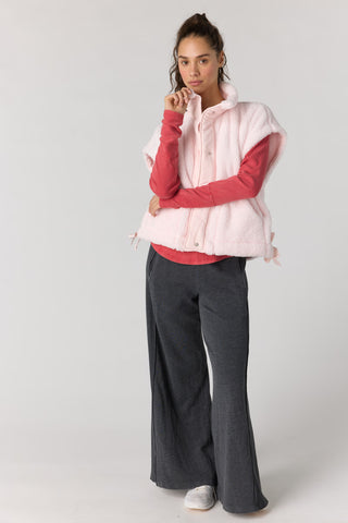 A model stands against a light gray studio background wearing a pale pink sleeveless puffer vest over a coral pink long-sleeve top, paired with dark charcoal wide-leg pants and white sneakers.