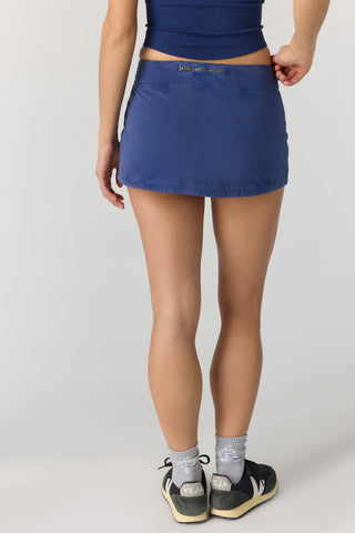 A person shown from the waist down wearing a blue athletic mini skort, a matching blue crop top, gray socks, and dark sneakers against a light gray studio background.