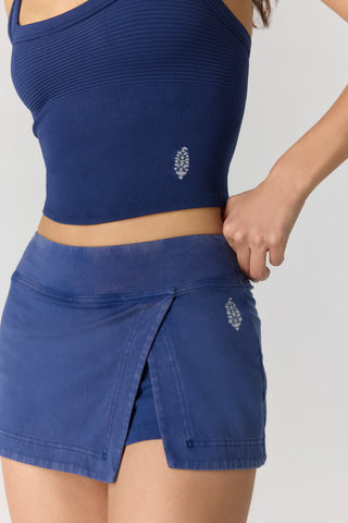 A close-up of a model wearing a blue ribbed crop top and matching blue wrap-style athletic skort with small silver leaf logos, against a light gray studio background.