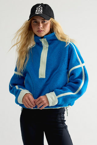 A woman wearing a bright blue sherpa fleece half-zip sweatshirt with white contrast panels and black pants, paired with a black baseball cap, against a plain light gray studio background.