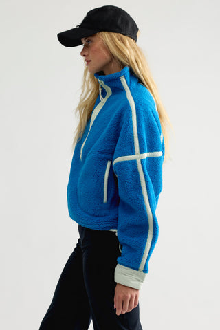 A woman in profile wearing a bright blue fleece pullover with white geometric stripe details, black pants, and a black baseball cap, photographed against a plain light gray studio background.