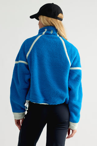 A person shown from the back wearing a bright blue fleece jacket with white contrast stitching and light gray ribbed cuffs, paired with black pants and a black baseball cap against a plain light gray studio background.
