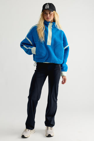 A blonde woman wearing a bright blue sherpa fleece quarter-zip jacket with light green contrast panels and a black baseball cap, photographed against a plain light gray background.