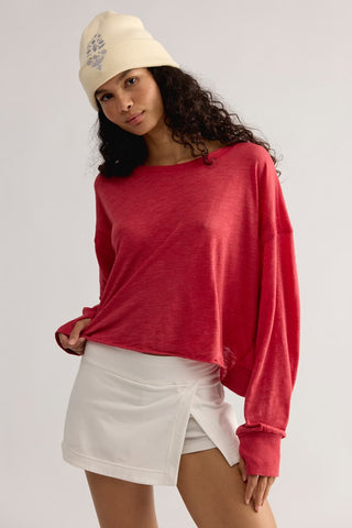 A woman wearing a faded red long-sleeve crewneck t-shirt, a white wrap-style mini skirt, and a cream-colored beanie with a gray graphic, posed against a light gray studio background.