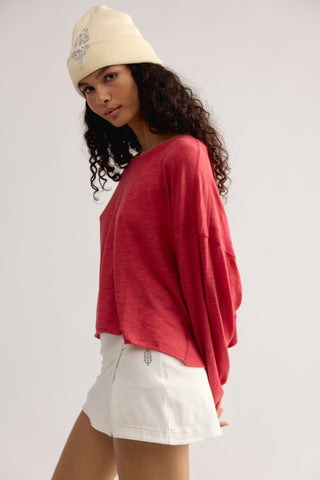 A woman wearing a loose-fitting, faded red long-sleeve t-shirt, white shorts, and a cream beanie with a blue floral design, photographed against a light gray studio background.