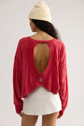 A woman shown from the back wearing a red long-sleeve cropped top with a large open back cutout, paired with a white skirt and cream beanie, against a light gray studio background.